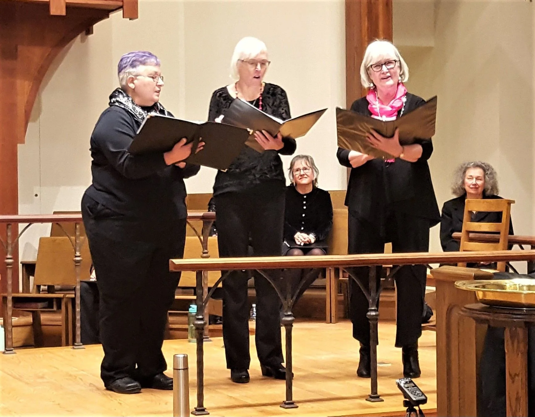 Three women are standing and singing with sheet music in a room with wooden details. Two women are seated behind them, one with curly gray hair and another with short gray hair and glasses. The women are dressed in black with accessories, and the set