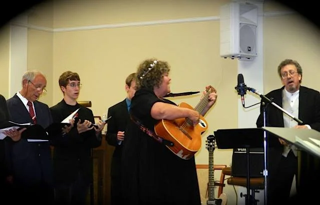 A group of six people in a room, some holding sheet music, with a woman singing and playing an acoustic guitar, and a man conducting or leading them, with musical equipment around.