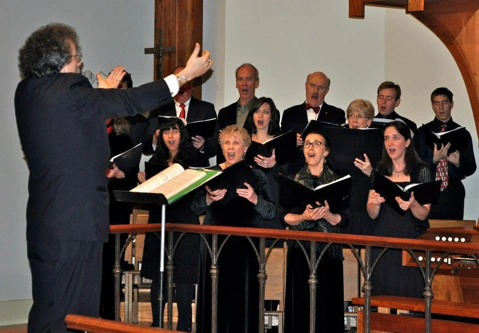 A choir of men and women singing under the direction of a conductor in a church or concert hall.
