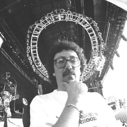 A person with glasses and a mustache in front of a large illuminated heart-shaped clock at night.