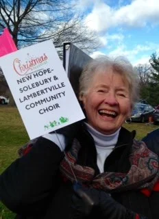 An elderly woman smiling and holding a sign that reads 'Christmas NEW HOPE - SOLEBURY & LAMBERTVILLE COMMUNITY CHOIR' outdoors on a cloudy day.