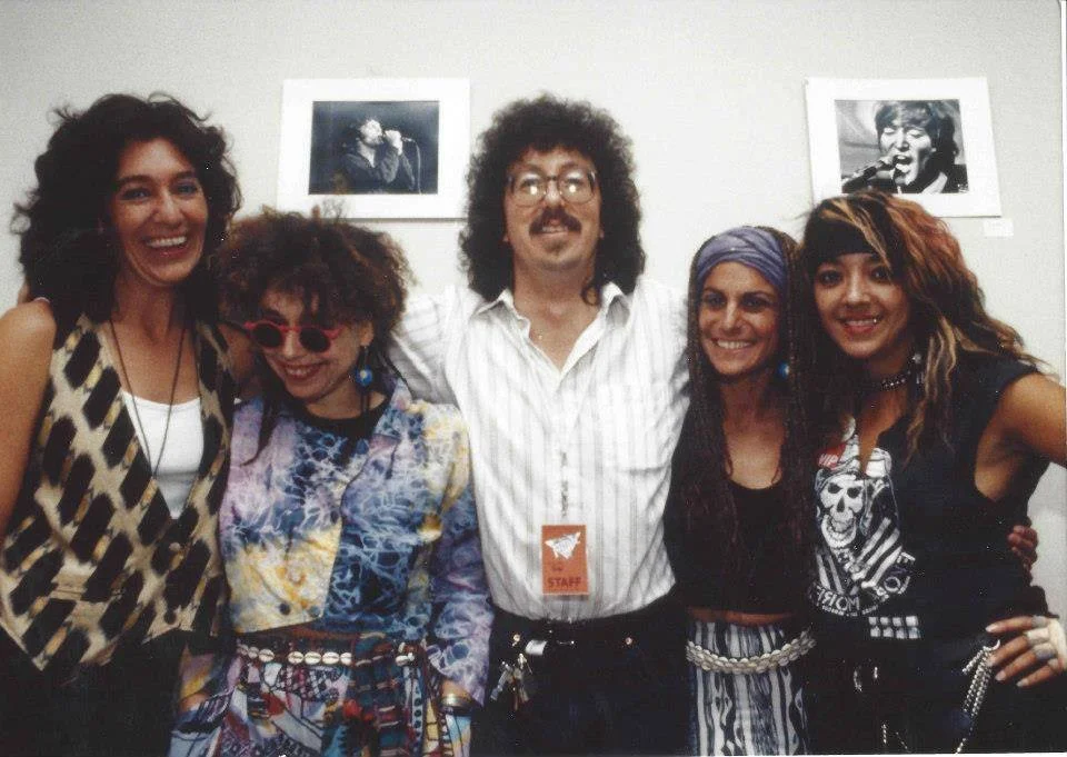 Group of five people with 80s fashion, smiling, standing in front of framed photos on a white wall.