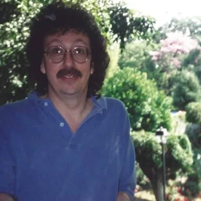 Man with curly hair, mustache, and glasses wearing a blue shirt standing outdoors with trees and bushes in the background.