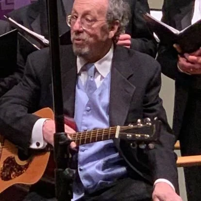 Old man with gray hair and beard playing an acoustic guitar in a formal setting, surrounded by choir members holding sheet music.