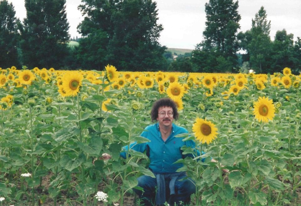 Man with curly hair and glasses crouching in a field of blooming sunflowers with deciduous trees in the background.