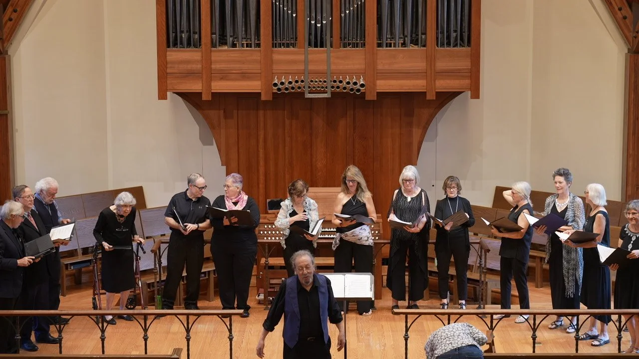 A choir of diverse adults standing on stage in a church, holding sheet music, with a conductor in front and a large wooden organ behind them.