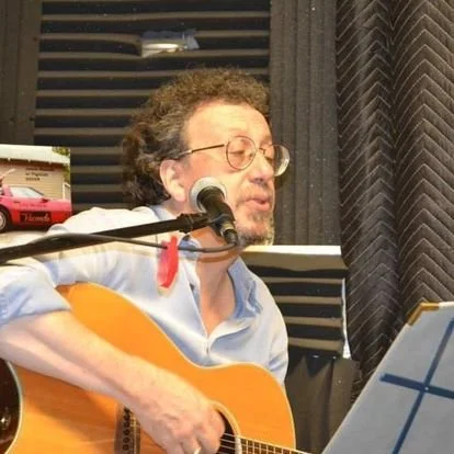 A man with curly hair, glasses, and a beard singing into a microphone while playing an acoustic guitar in a recording studio.