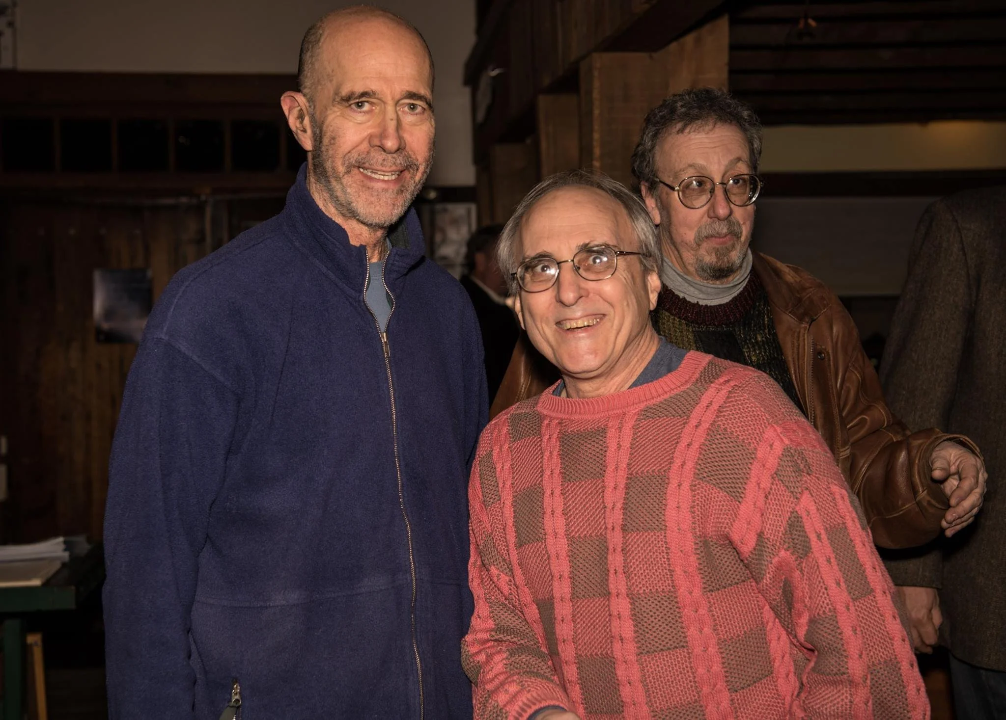 Three men are standing together indoors. The man on the left is wearing a blue zip-up jacket and has a beard. The man in the middle is wearing glasses and a red and gray patterned sweater, smiling. The man on the right is wearing glasses, a dark swea
