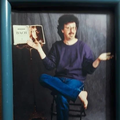Person sitting cross-legged on a chair holding a book about Bach music, with a dark background.