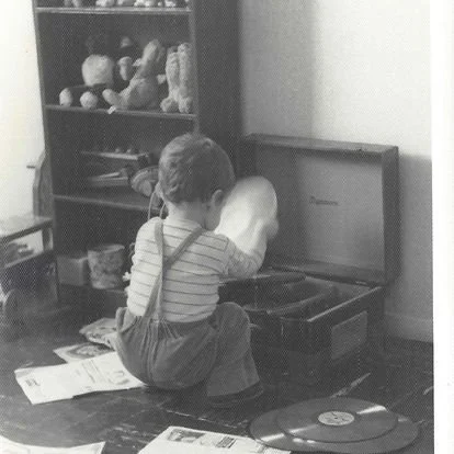 A young child sitting on the floor and opening a large, vintage-style trunk or box in a room with shelves filled with toys and items.