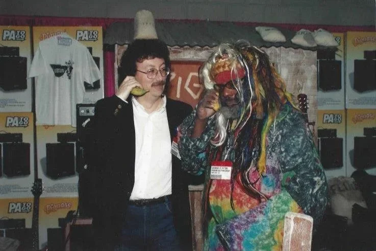 Two people dressed as Steve Jobs and a colorful hippie, standing in an indoor setting with display booths and T-shirts in the background.