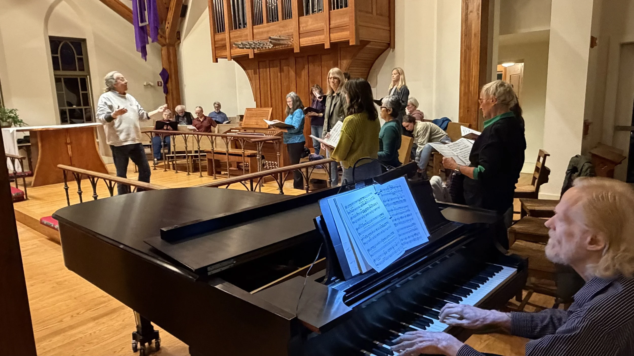 A group of people singing in a church, led by a man in a white shirt. A piano with sheet music in the foreground. The church has wooden accents and a balcony above.