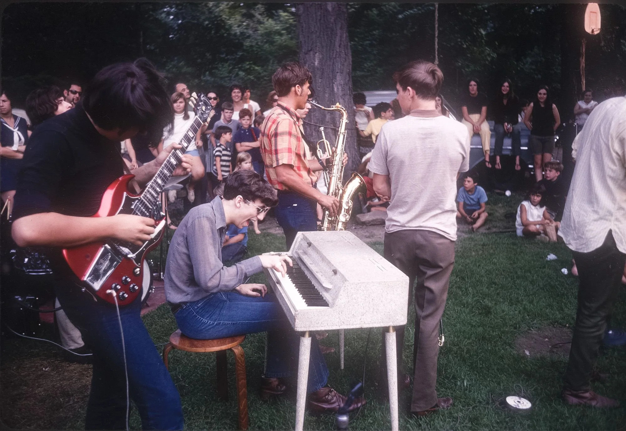 A group of musicians performing outdoors in a park, with a crowd of people watching. The band includes a guitarist, a keyboard player, and a saxophonist. The audience consists of children and adults standing and sitting on the grass.
