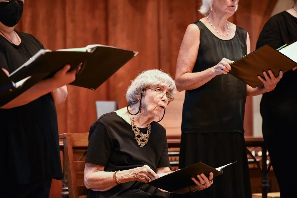 An elderly woman in black seated while singing in a choir, holding a black folder, with other choir members standing and singing around her in a room with wood panel walls.
