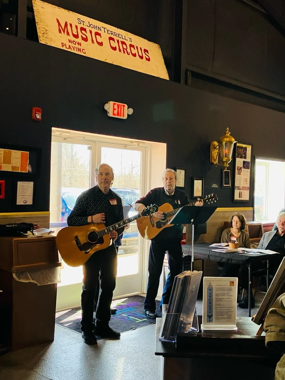 Two men playing guitars in a restaurant with a music-themed sign overhead, an exit door behind them, and seated audience members.