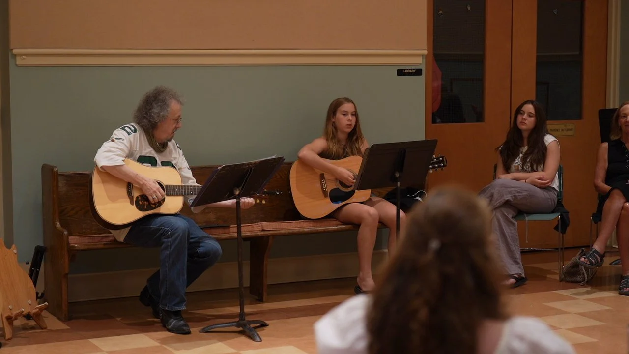 Three women sitting in a room, two of them playing acoustic guitars, with an audience member visible in the foreground.