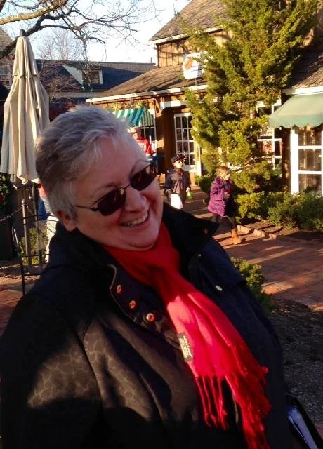 A smiling woman with short gray hair, wearing sunglasses, a red scarf, and a dark jacket, standing outdoors in front of a brick walkway, with other people and buildings in the background.