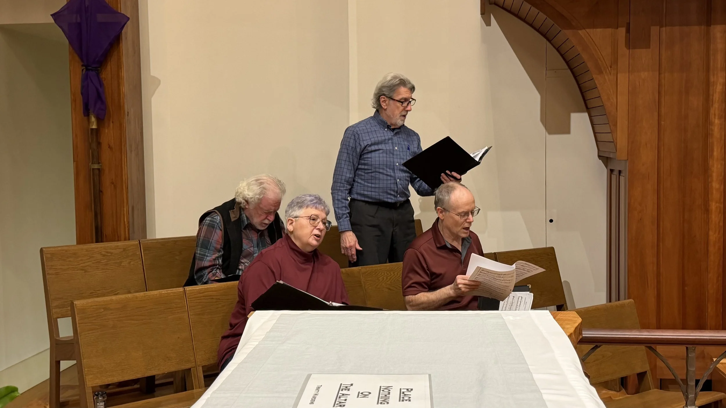 Group of five elderly people, three men and two women, seated and standing at a church or community hall, singing from printed sheets of music or hymnals.
