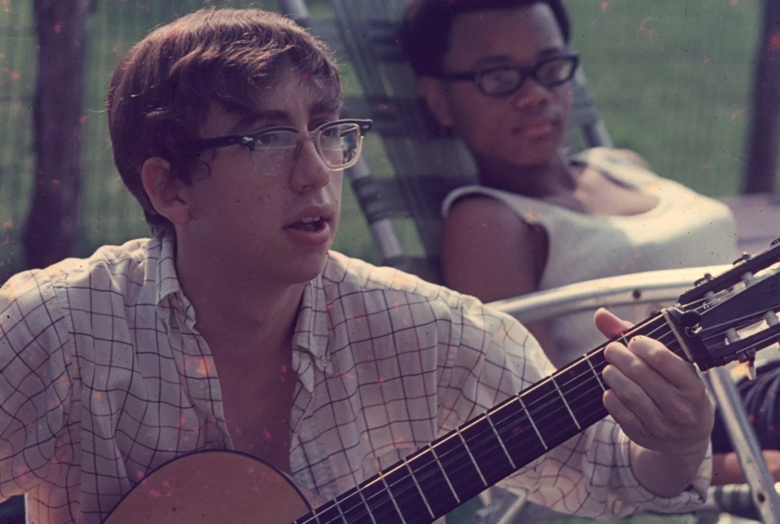 Two young people wearing glasses relaxing outdoors, one playing a guitar, sitting on lounge chairs.