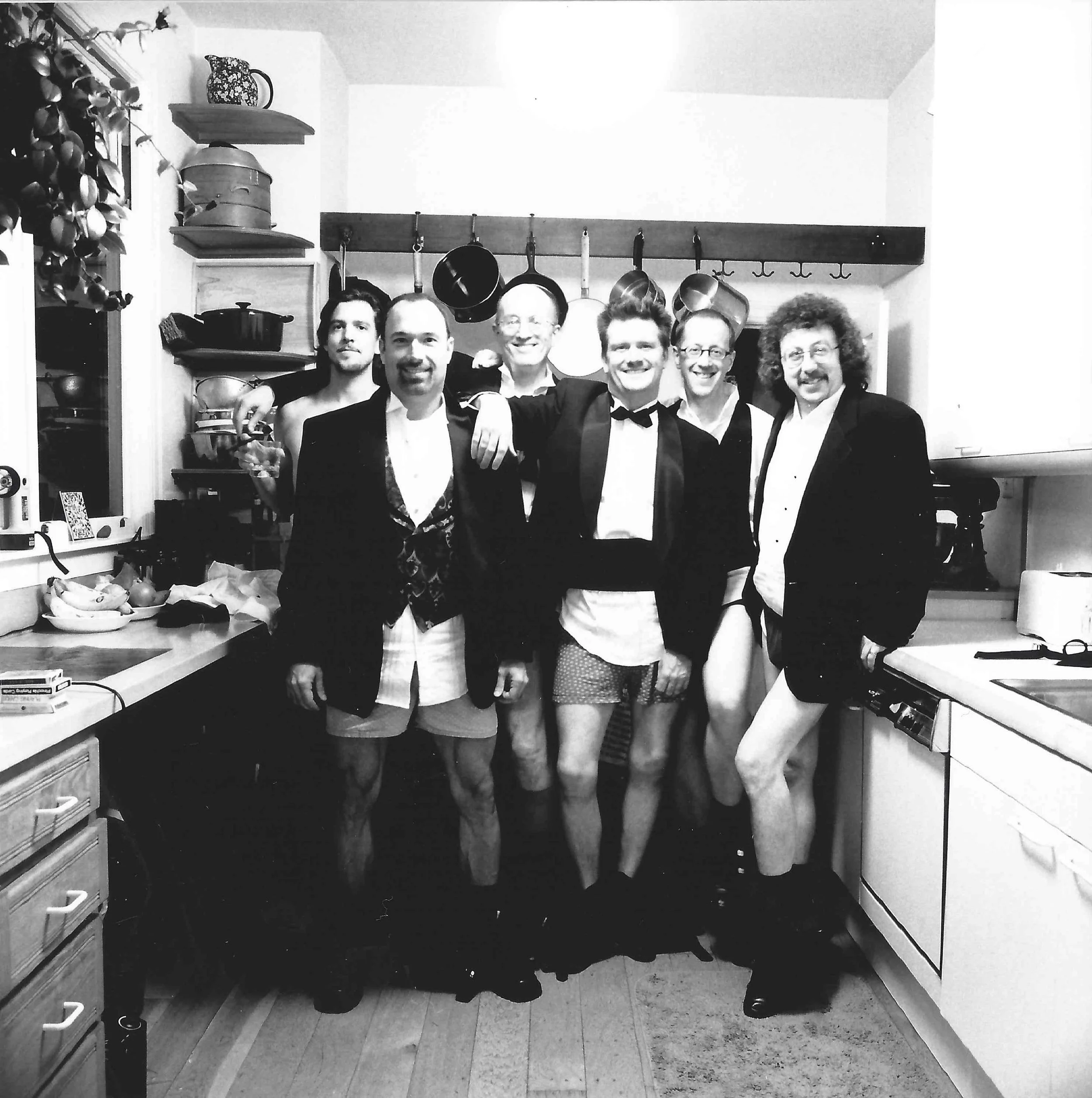 A group of six men standing together in a kitchen, smiling and dressed in formal attire, with some wearing tuxedos and bow ties, and others in suits. They have their arms around each other.