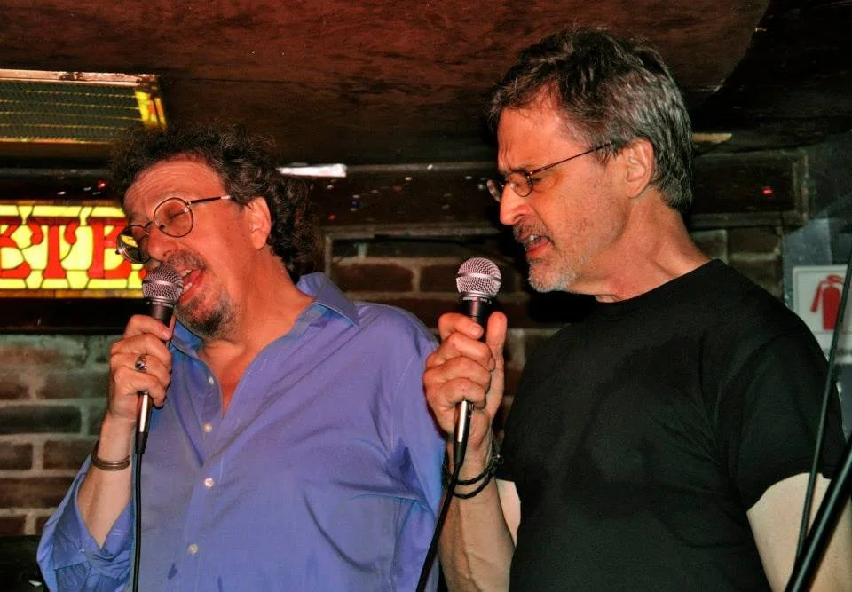 Two men singing into microphones at a karaoke or open mic night, in a dimly lit bar or club with brick walls.
