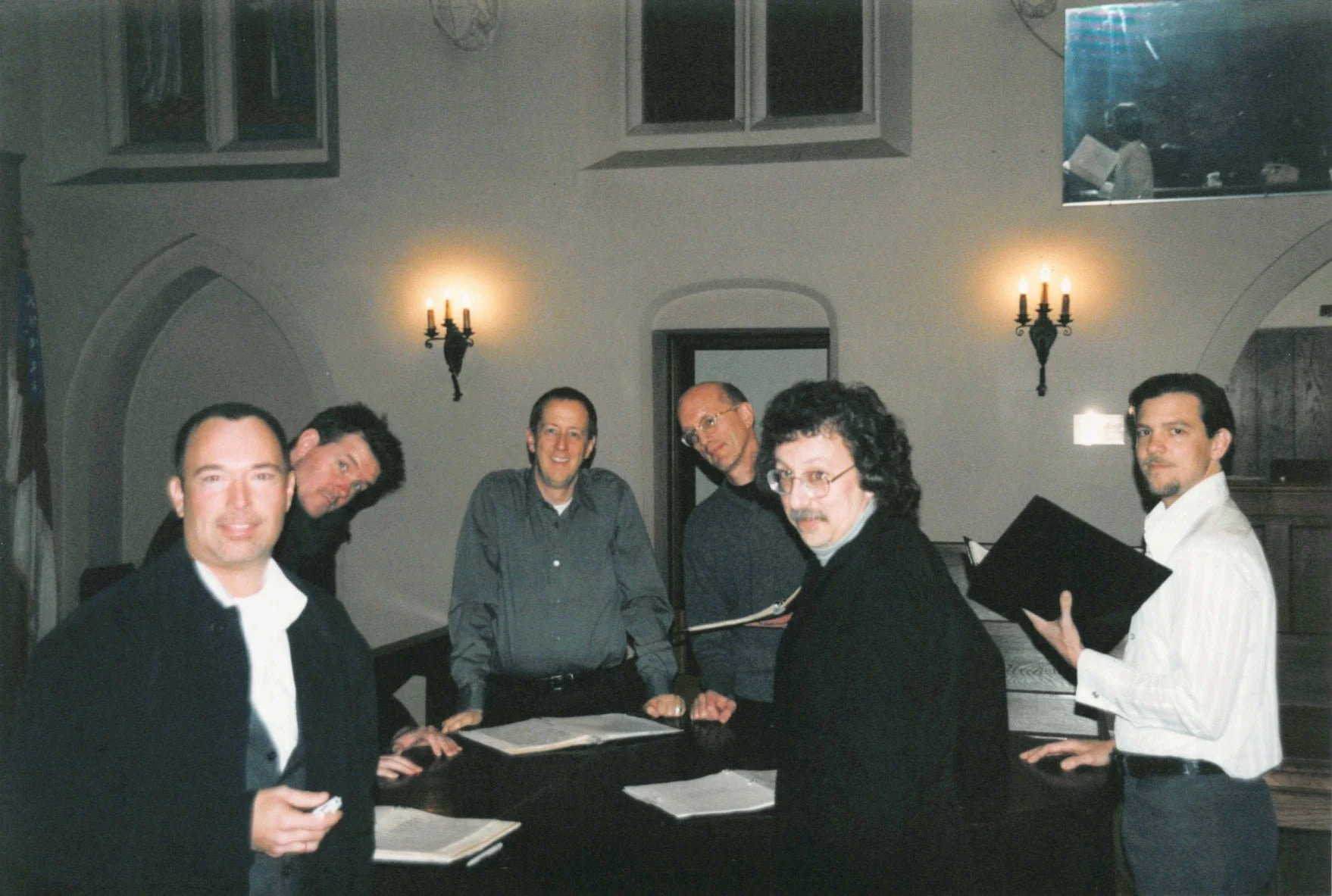 A group of six men standing around a table in a room with beige walls, two windows, wall sconces, and a mirror. Some are smiling, one is holding a black music folder.