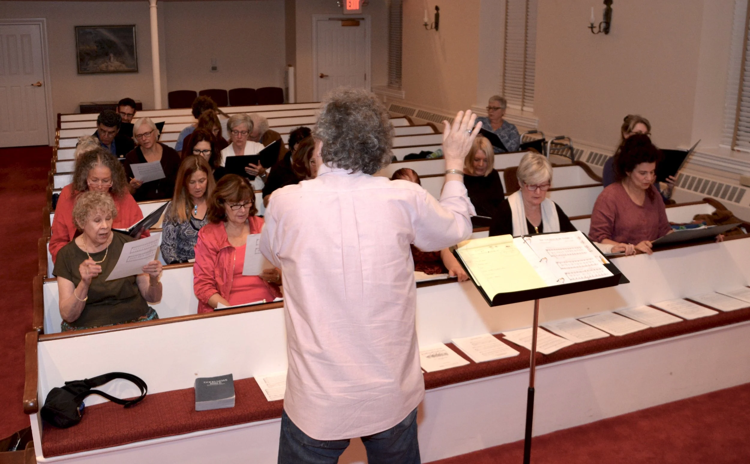 A group of people, mostly women, sitting in pews inside a church or chapel, practicing choral singing, with a conductor standing in front of them, leading and holding music sheets. The room has white walls, wooden pews, and several windows with blind