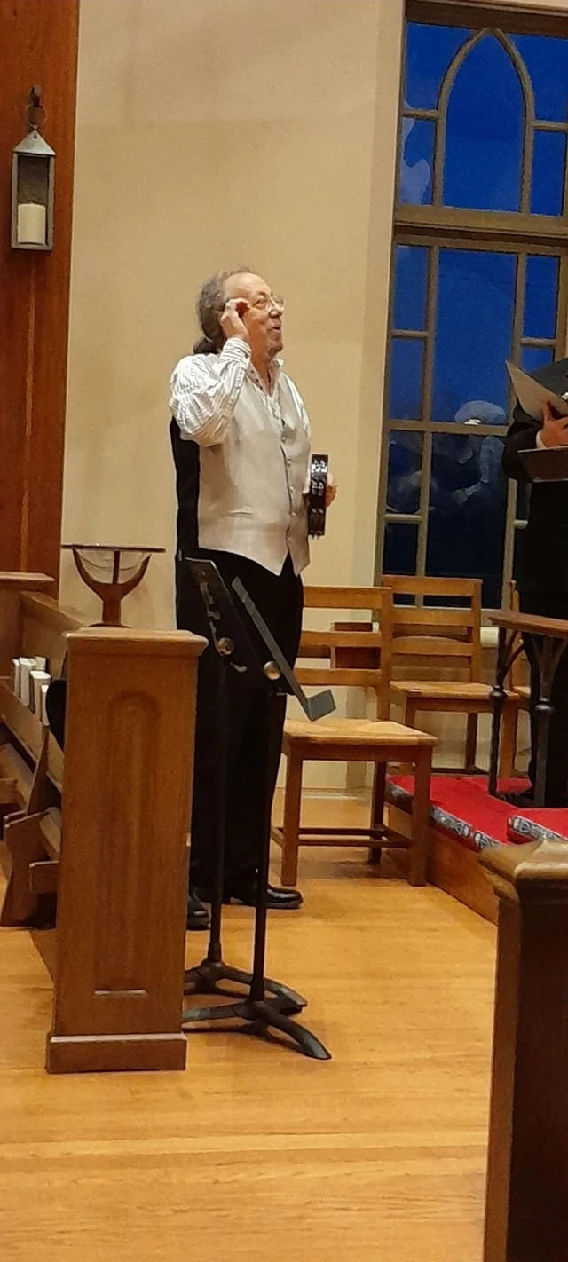 An elderly woman standing inside a church, holding a phone and wearing glasses, with a large window in the background and wooden pews around her.