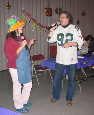 A woman wearing a colorful clown wig and bright clothing holding a microphone, and a man in a football jersey holding a microphone, standing in front of a decorated background featuring hanging balloons and a banner that says 'Happy Birthday'.