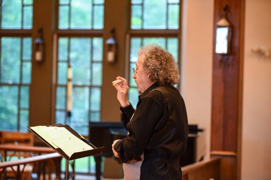 A man with curly gray hair, glasses, wearing a black shirt, standing at a music stand inside a church or chapel, with tall windows and wooden accents.