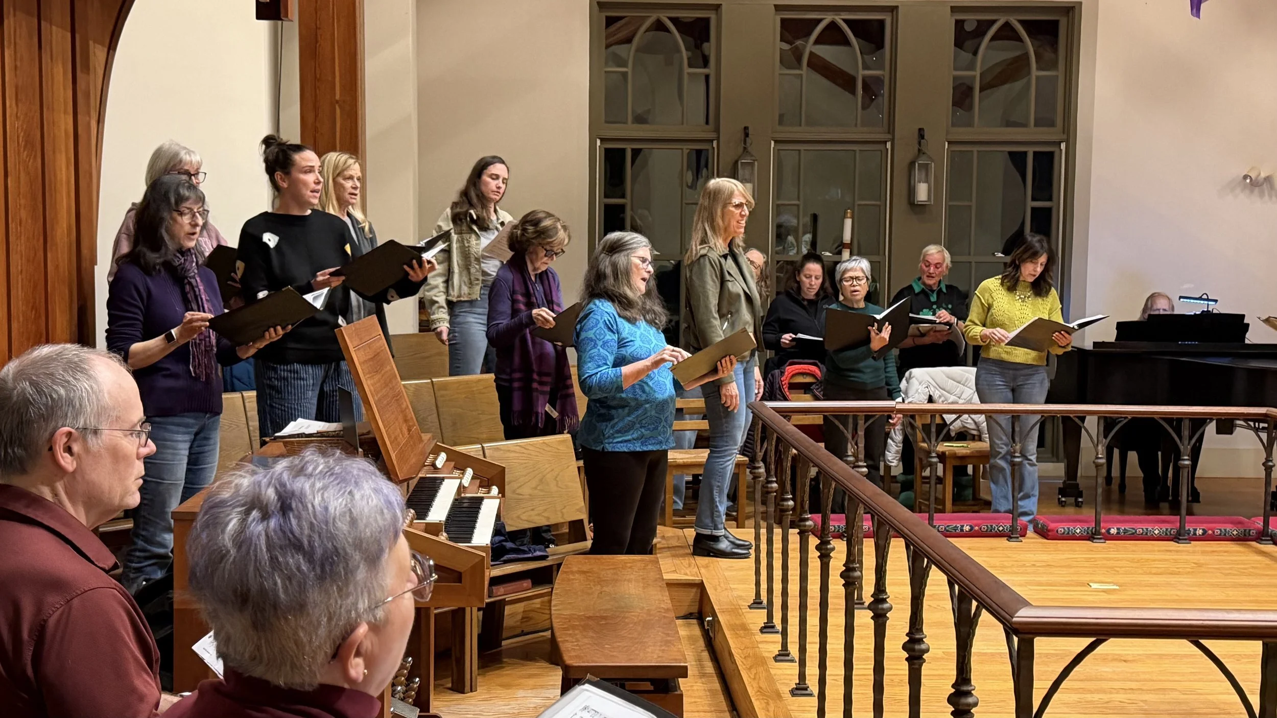 A choir of men and women singing in a church or concert hall, with a pianist in the foreground and audience members seated in front.