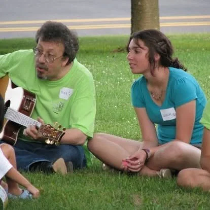A man and a woman sitting on grass outdoors with a tree in the background. The man is playing guitar and the woman is listening.