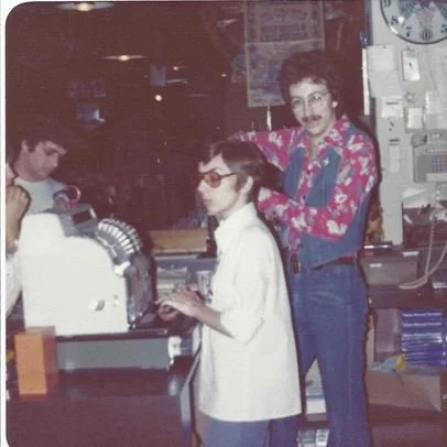 A woman with curly hair and glasses, standing behind two children at a laboratory bench, smiling and wearing a colorful shirt with a floral pattern, in a vintage science classroom.