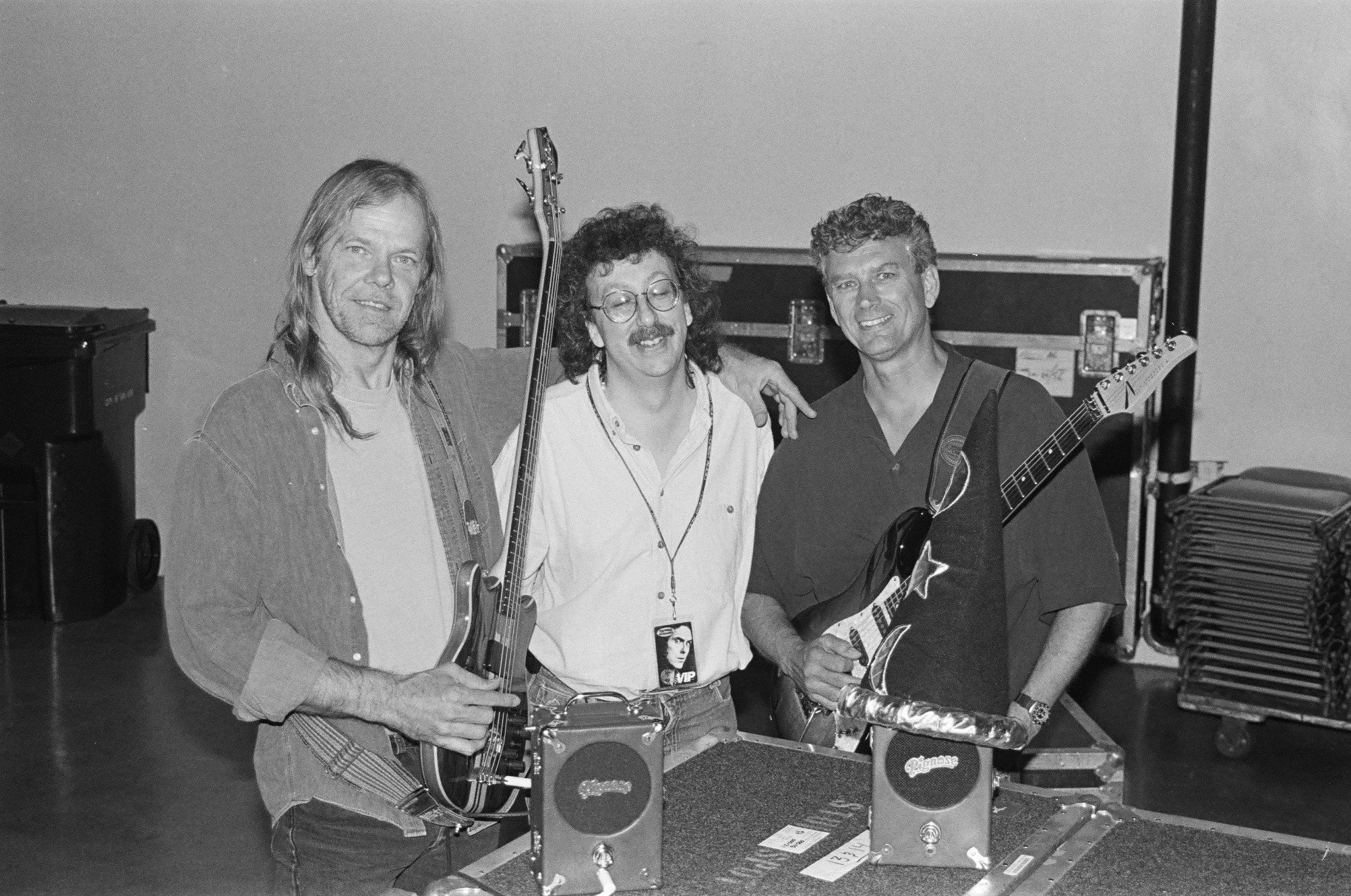 Black and white photo of three men in a music studio, two are holding guitars, one is wearing a VIP badge, and there are music equipment and cases in the background.