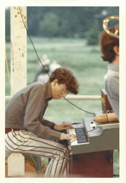 A person with curly hair playing a piano outdoors near a body of water, with a woman wearing a hat partially visible on the right.