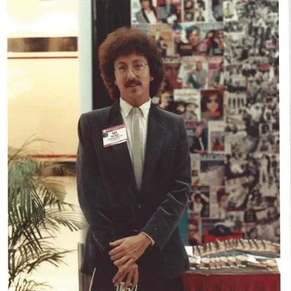 Man with curly hair and glasses in a suit standing indoors near a collage of photos.