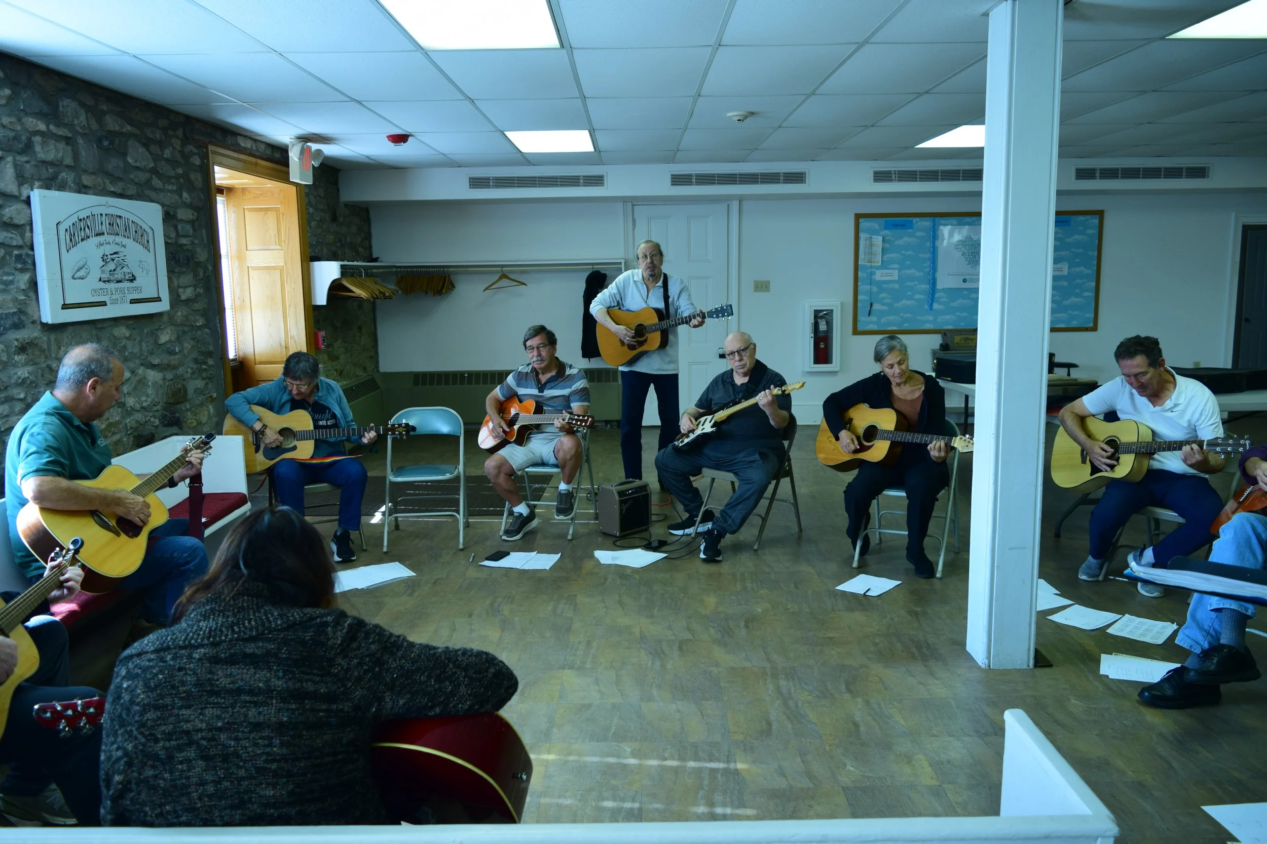 Group of people playing guitars in a room, with one person standing with a guitar, others seated, and sheet music on the floor. The room has a stone wall, a sign on the wall, and a bulletin board.