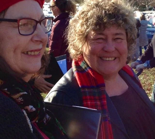 Two women smiling outdoors during a gathering or event, one wearing glasses and a Santa hat, the other with curly hair and a red plaid scarf.