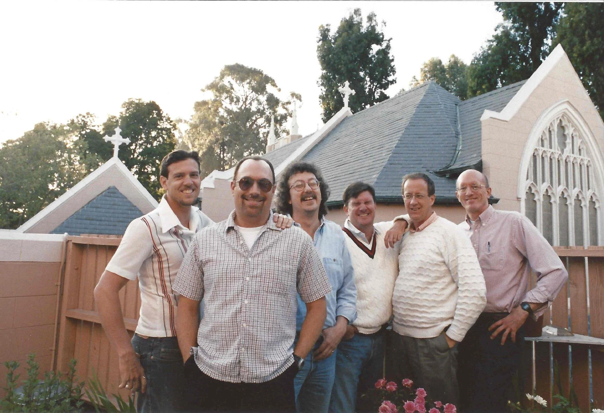 A group of seven men standing together outdoors in front of a church with a gray roof and stained glass window, smiling at the camera, with trees and a garden with pink flowers in the background.