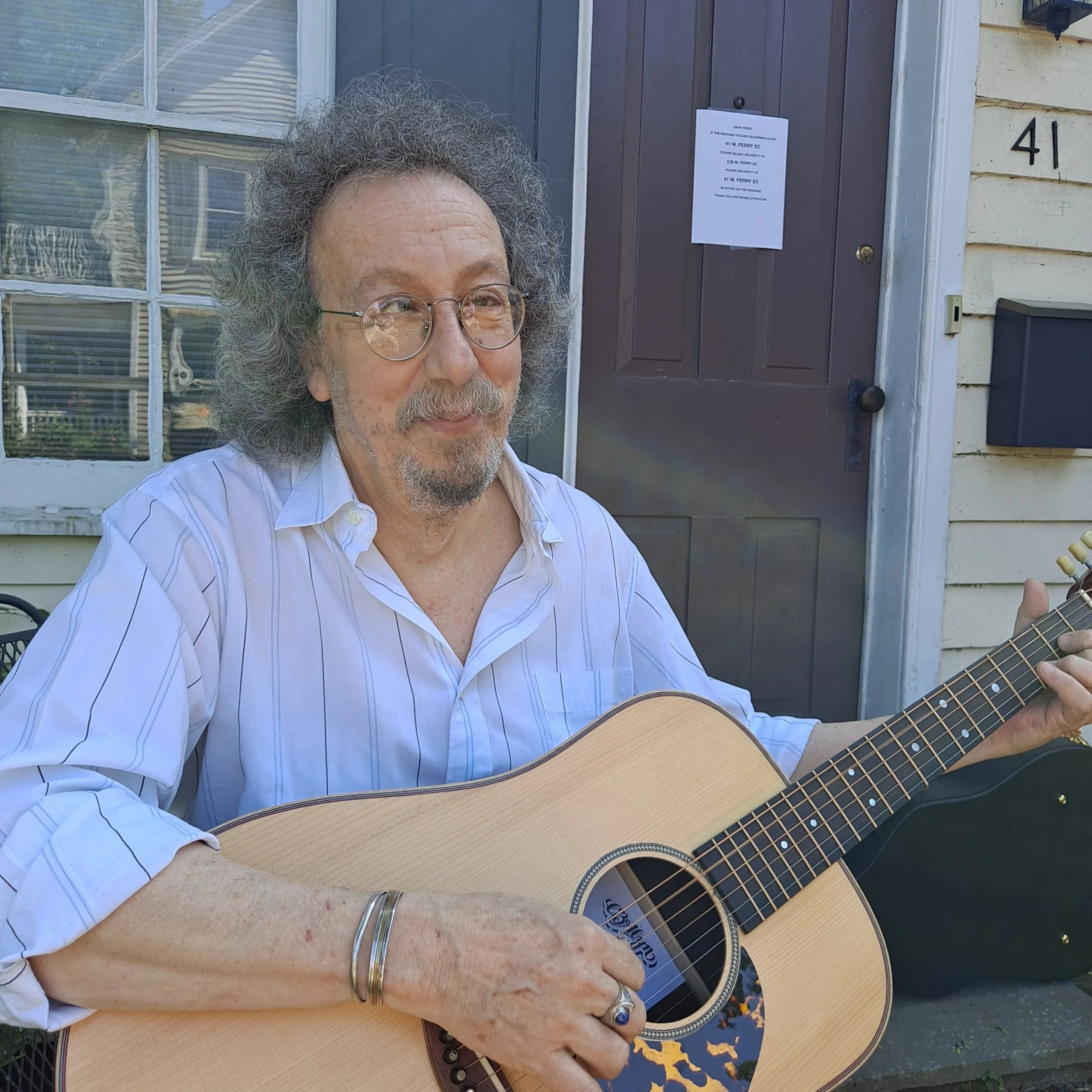 A man with curly gray hair and glasses playing an acoustic guitar outdoors in front of a house with the number 41 on the wall.