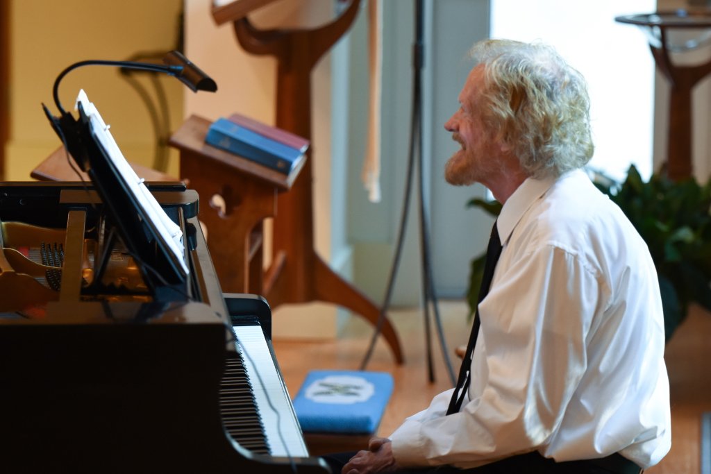 An elderly man with white curly hair and a beard, dressed in a white shirt and black tie, sitting at a piano and smiling.