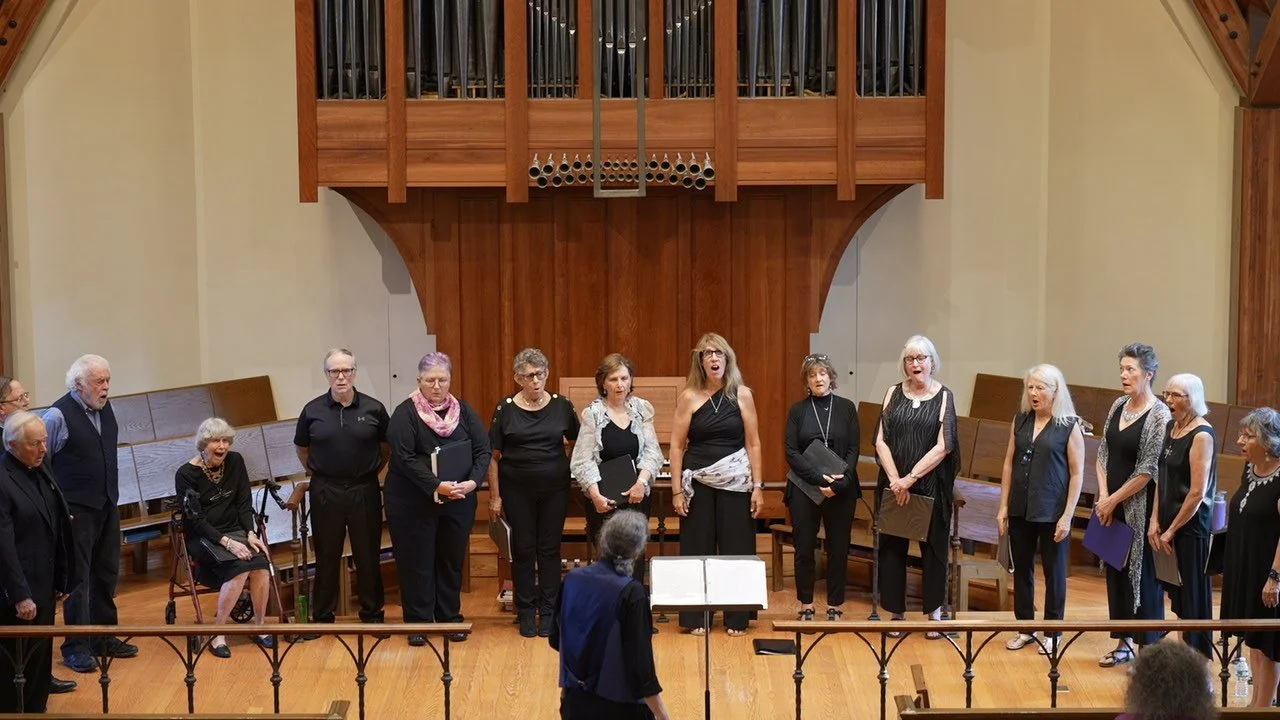 A choir of men and women standing on a stage inside a church, singing while a conductor leads them.
