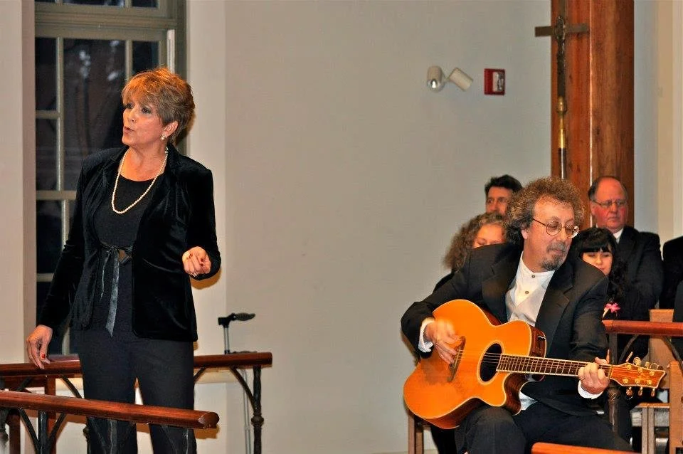 A woman singing or speaking with a man playing an acoustic guitar during an indoor event, with audience members in the background.