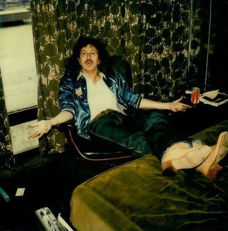 A man with curly hair and a mustache relaxing on a bed with a green cover, wearing a shiny blue jacket over a white shirt, with a soda can in his left hand, in a room with dark floral curtains and a window.