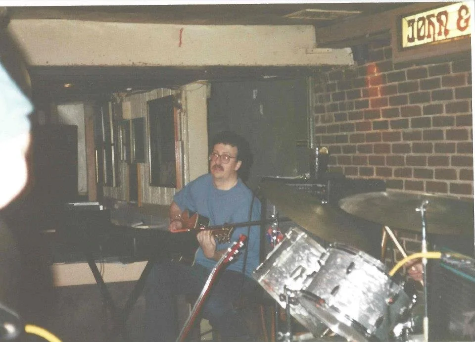 A man seated at a keyboard playing an acoustic guitar, surrounded by drum equipment, in a dimly lit bar or music venue with brick walls.