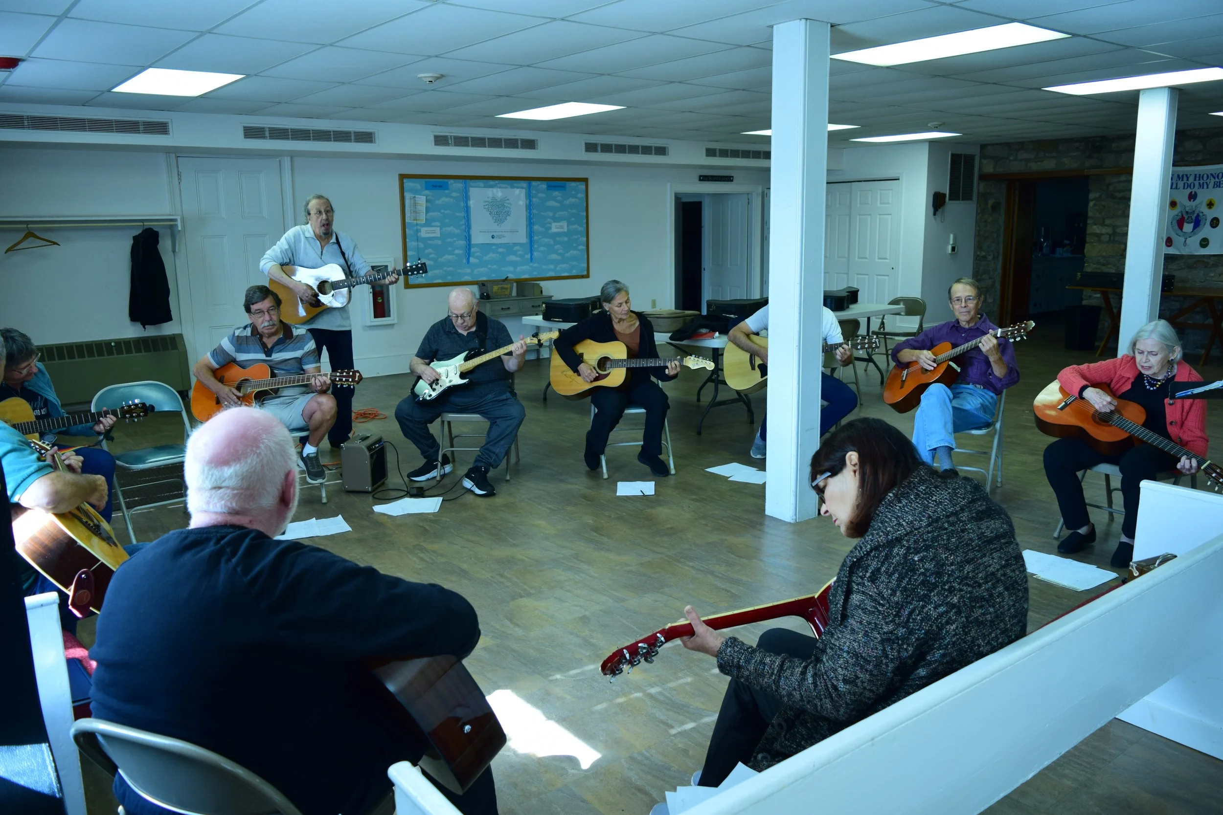 A group of elderly people playing guitars and singing in a room, with a person standing and singing at the front.