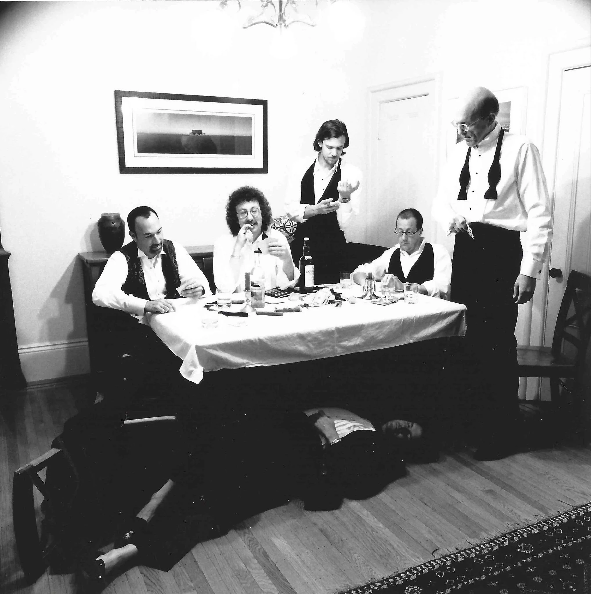 A group of people gathered around a dining table in a room with hardwood floors and artwork on the wall, some standing and some sitting, engaged in conversation and eating.
