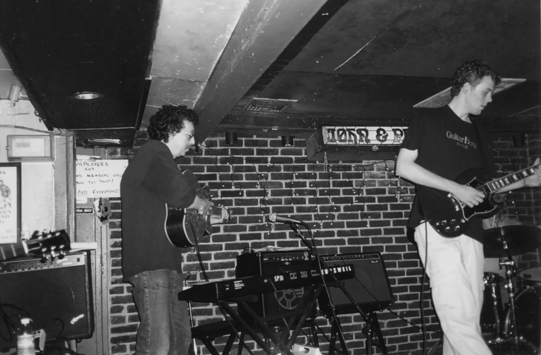 Two musicians playing guitars on a brick-walled stage in a bar or club, with a sign reading 'JONES & P' in the background, and a small amplifier and equipment nearby.
