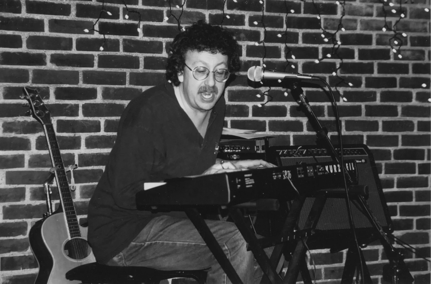 A man with curly hair and glasses playing a keyboard in front of a brick wall, with a guitar standing nearby.