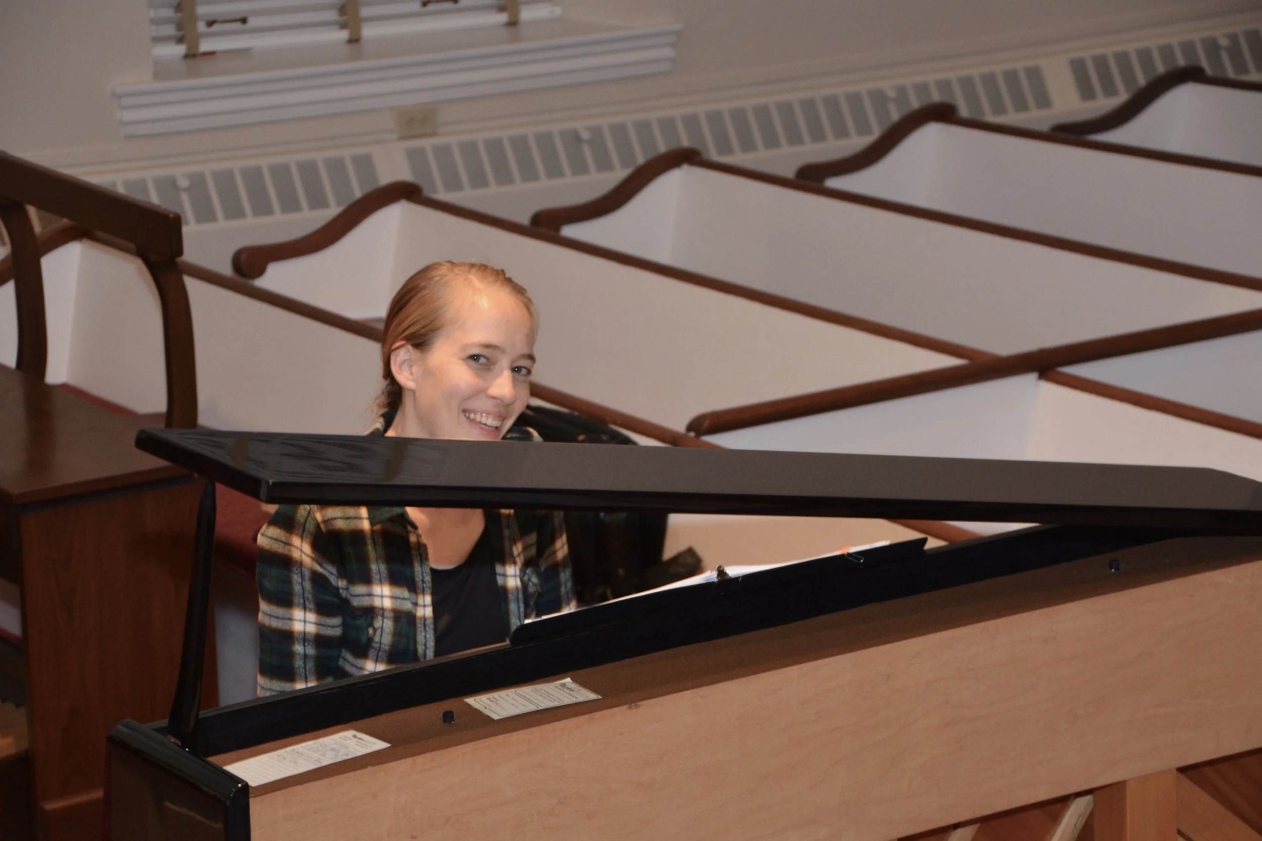 A woman with reddish hair smiling while sitting at a black grand piano in a room with wooden floors and empty church pews.